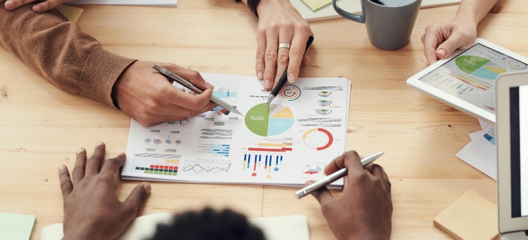 A team of professionals collaborating over printed charts and graphs at a table, discussing marketing strategies and performance metrics to understand how much should moving companies invest in marketing.