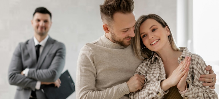 A happy couple embracing in their new home, with a moving manager standing in the background.