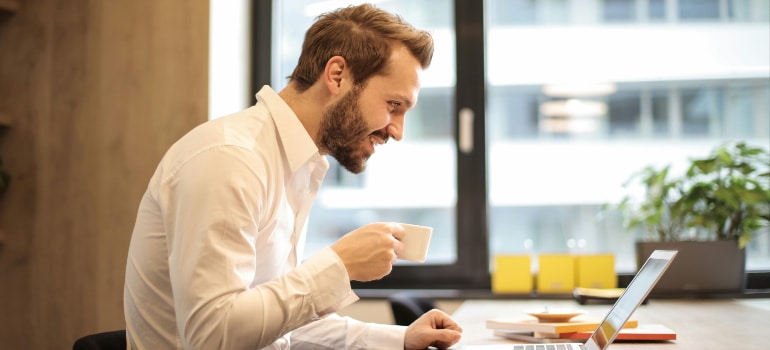 Man enjoying coffee while working remotely on his laptop in a modern office. Highlights remote work and moving trends, showing work flexibility.