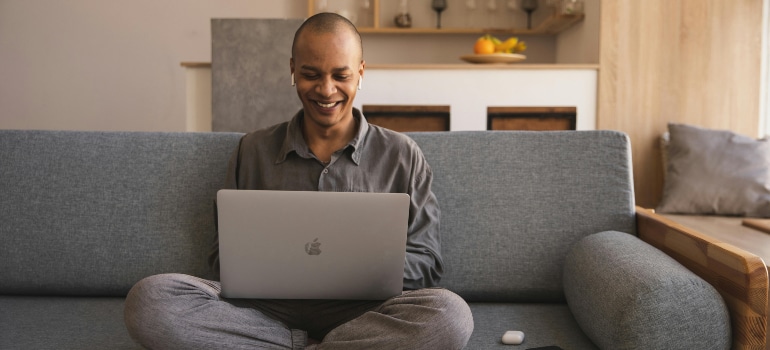 Man sitting on a couch, smiling while working remotely on his laptop at home. Represents remote work and moving trends, illustrating home office comfort.