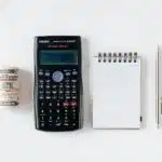 Money, a calculator, a note pad, and a pen on a white surface