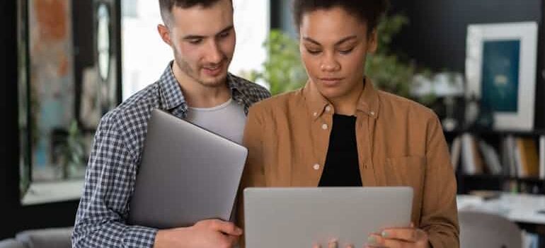 A young man holding a white tablet computer standing next to a woman holding her tablet while showing him how AI helps movers write better emails.