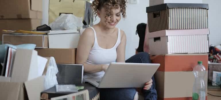 A woman sitting on the floor surrounded by packed boxes, using her laptop to connect with professional movers and plan her relocation.