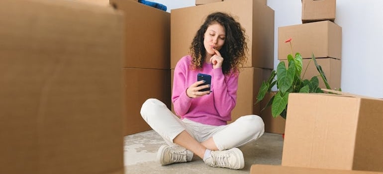 A person sitting on the floor, surround by boxes, while looking at their phone