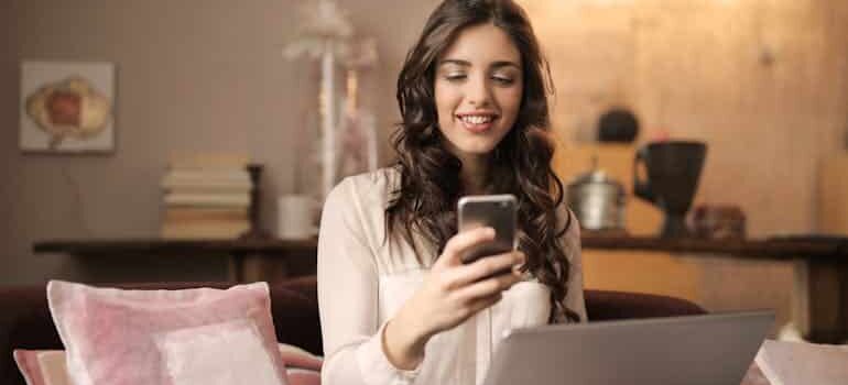 Person using a smartphone while viewing a laptop in a cozy living room, reviewing online business data and digital marketing performance.